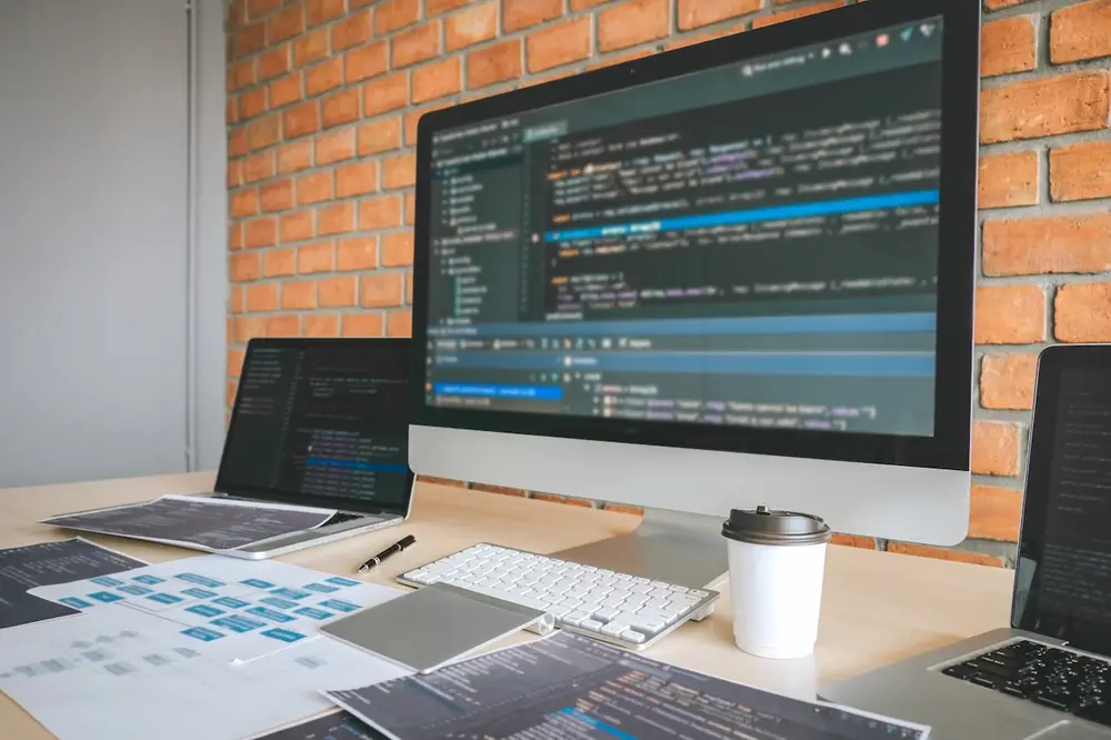 Programmeur au bureau travaillant sur un grand écran affichant du code source, avec ordinateur portable, clavier, documents et tasse à café sur un bureau en bois contre un mur en briques rouges.